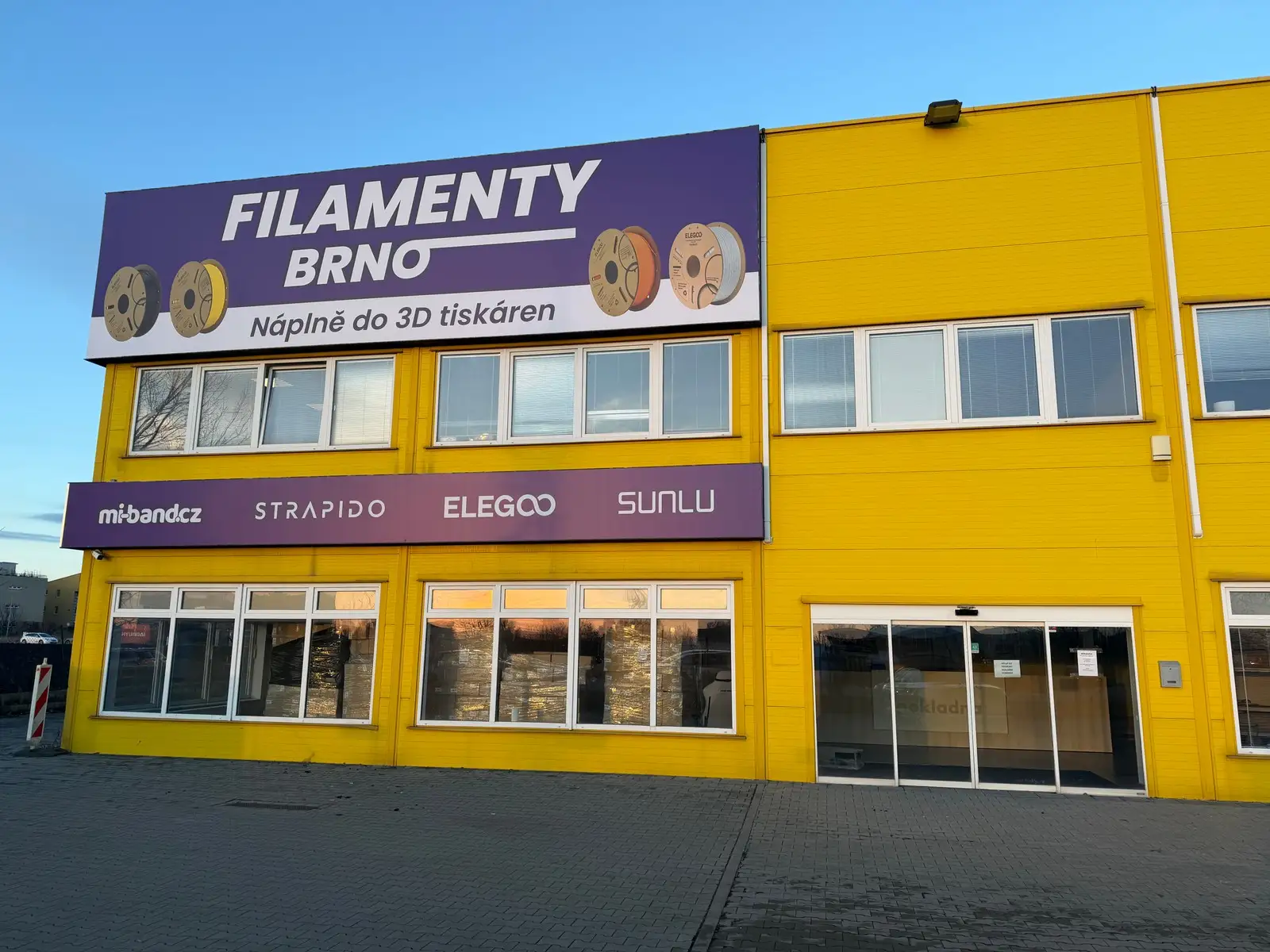 Žlutá budova prodejny FilamentyBrno s označením nad výlohami a vstupem vpravo